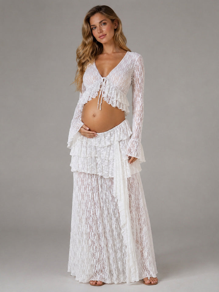 A pregnant woman modeling a white lace maternity two-piece set consisting of a long-sleeve tie-front crop top and a matching maxi skirt, photographed against a neutral gray studio background.
