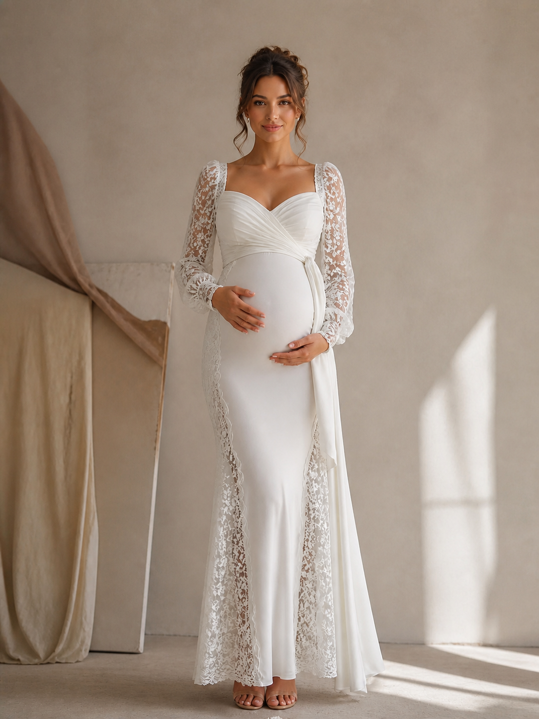 A pregnant woman wearing a white maternity gown with long lace sleeves and a fitted silhouette, standing in a softly lit studio with a neutral beige wall and draped fabric.