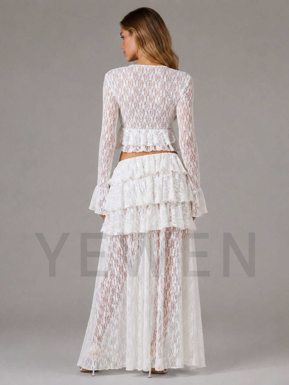 A woman shown from the back wearing a white lace two-piece outfit with a long-sleeve cropped top and a tiered ruffled skirt, photographed against a plain gray studio background.