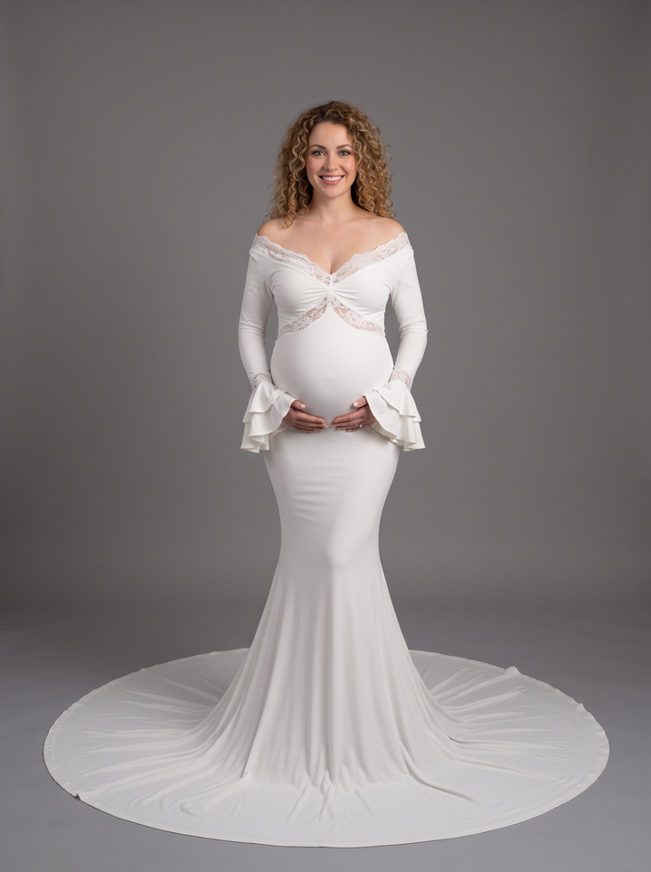 A pregnant woman wearing a white off-the-shoulder maternity gown with long flared sleeves, a lace-trimmed sweetheart bodice, and a fitted mermaid-style skirt with a long train, photographed against a plain gray studio background.