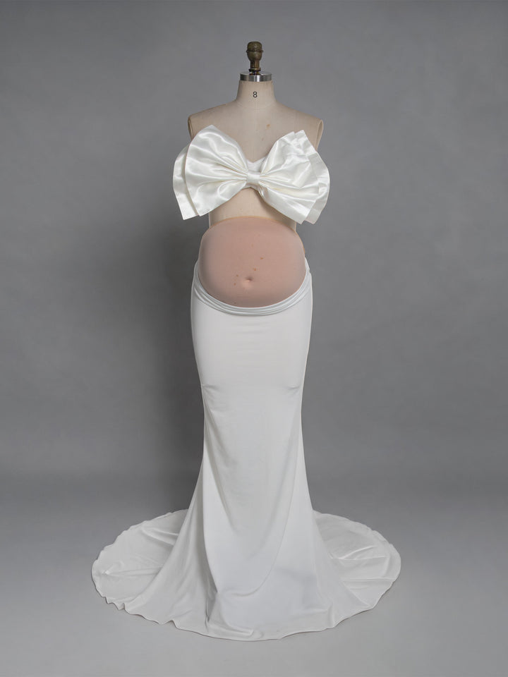 A white maternity dress displayed on a mannequin torso against a plain gray studio background. The dress has a large bow at the bust and a fitted, floor-length skirt with a slight train.