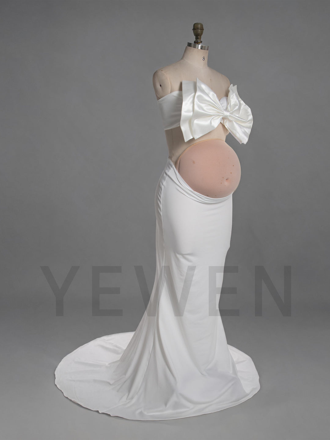 A white maternity gown displayed on a dress form mannequin against a gray studio background. The strapless gown features a large bow at the bust and a fitted silhouette that reveals a round belly cutout.