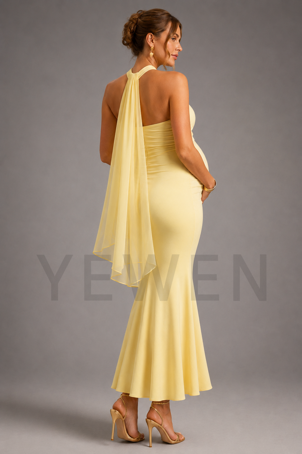 A pregnant woman is shown from the back wearing a pale yellow halter-neck maternity gown with a draped sheer overlay and a fitted silhouette, paired with nude high-heeled sandals against a gray studio background.