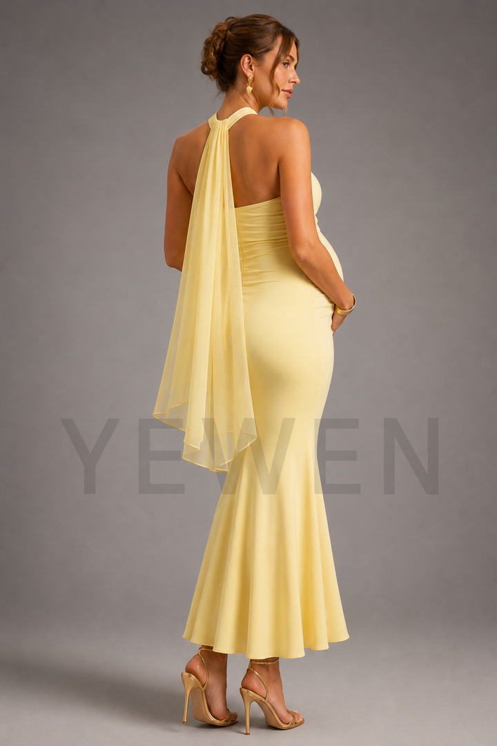 A pregnant woman is shown from the back wearing a pale yellow halter-neck maternity gown with a draped sheer overlay and a fitted silhouette, paired with nude high-heeled sandals against a gray studio background.
