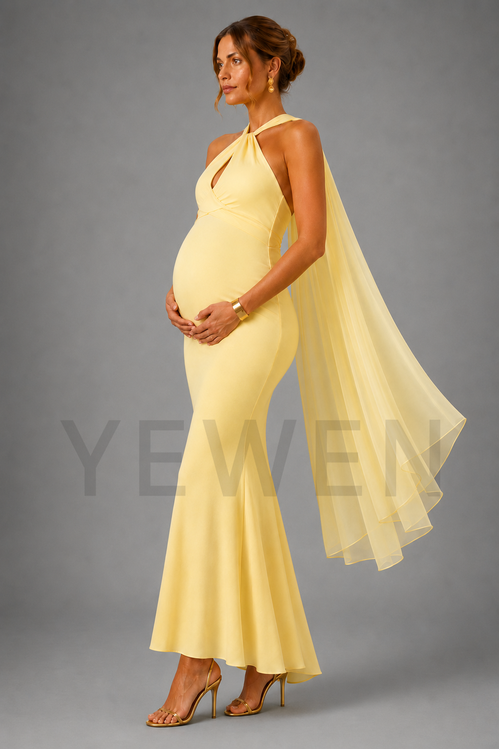 A pregnant woman modeling a pale yellow halter-neck maternity gown with a flowing sheer cape and gold heels, standing against a solid gray studio background.