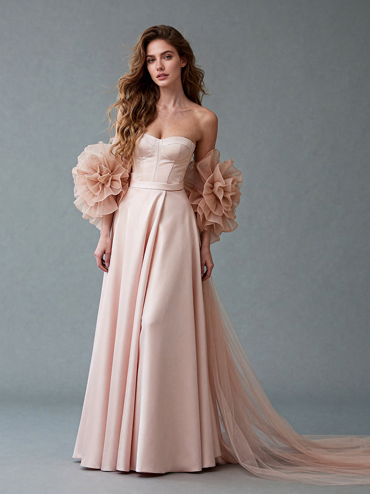 Full front view of a model wearing a blush strapless satin prom gown with a corset bodice and a 3D ruffle organza cape.