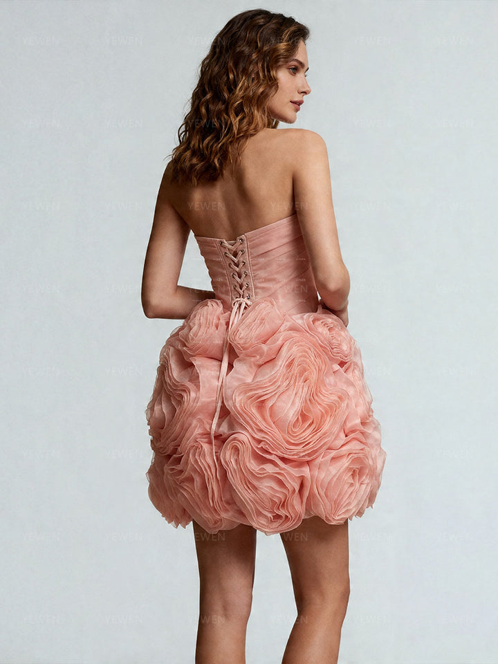 Back view of a model wearing a blush ruched 3D floral mini dress highlighting the adjustable lace-up corset back and voluminous bubble hem.