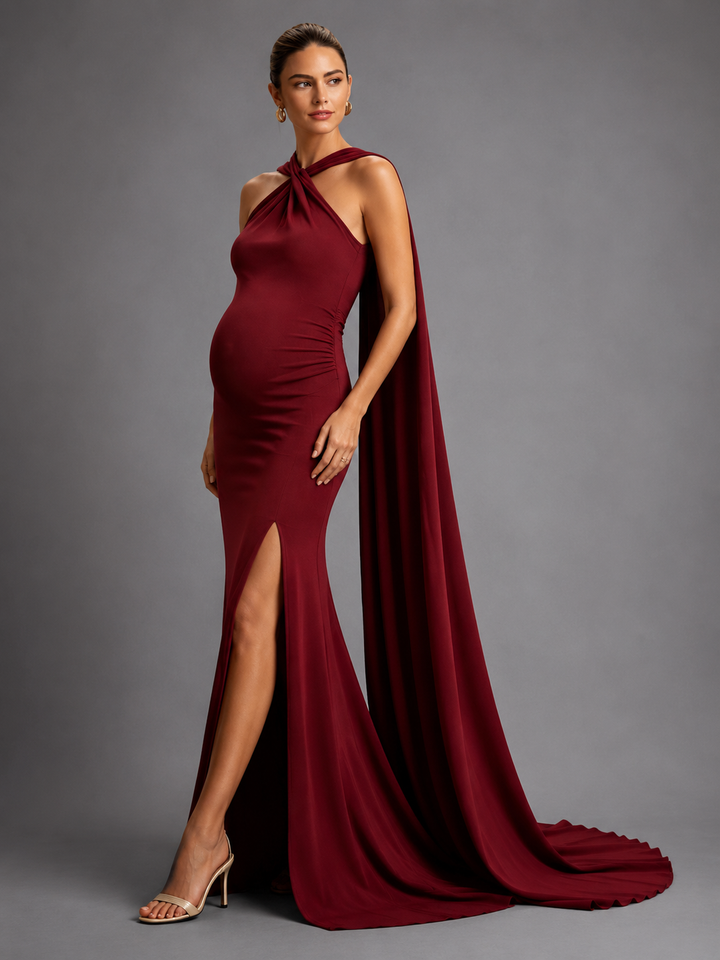 A pregnant model wearing a floor-length, deep burgundy halter gown with a high thigh slit and a dramatic draped train, posed against a solid gray studio background.