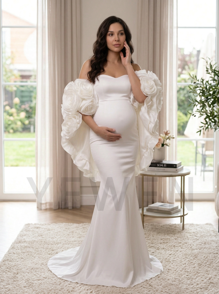 A pregnant woman stands indoors wearing an elegant off-the-shoulder white maternity gown with dramatic ruffled puff sleeves and a fitted mermaid silhouette. The room has large windows with beige curtains, a small side table, and a plush cream rug.