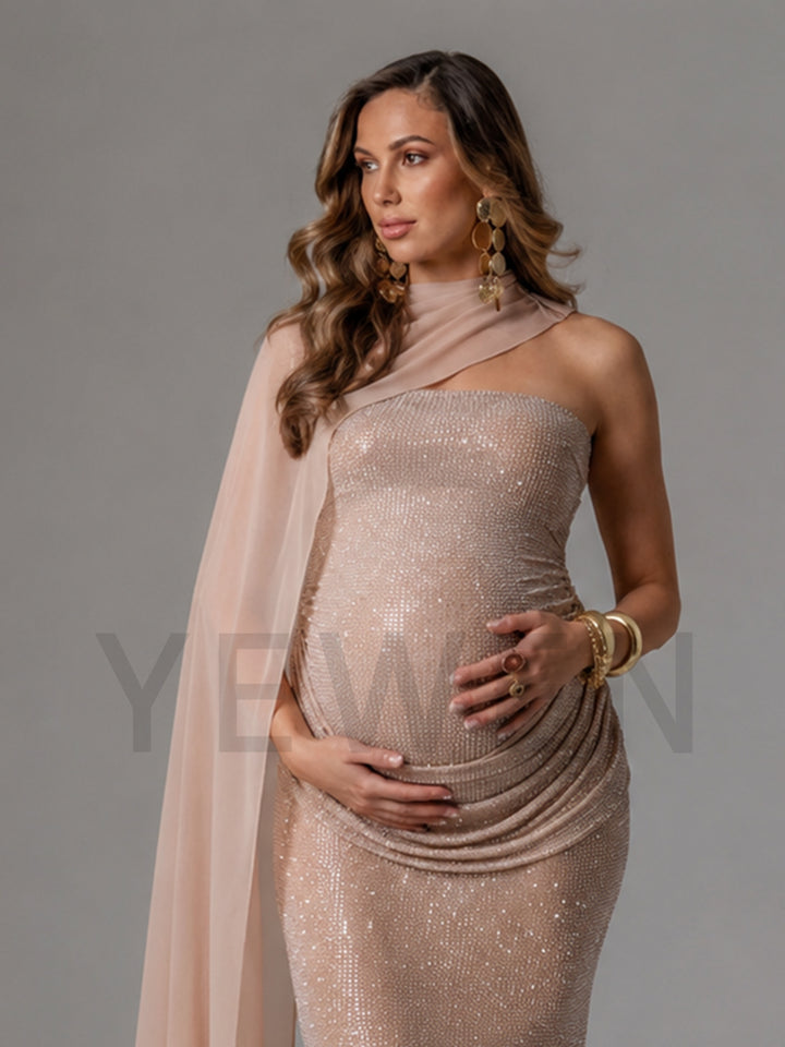 A pregnant woman wearing a shimmering champagne pink sequined one-shoulder gown with a matching sheer draped scarf, accessorized with gold jewelry, set against a solid gray studio background.