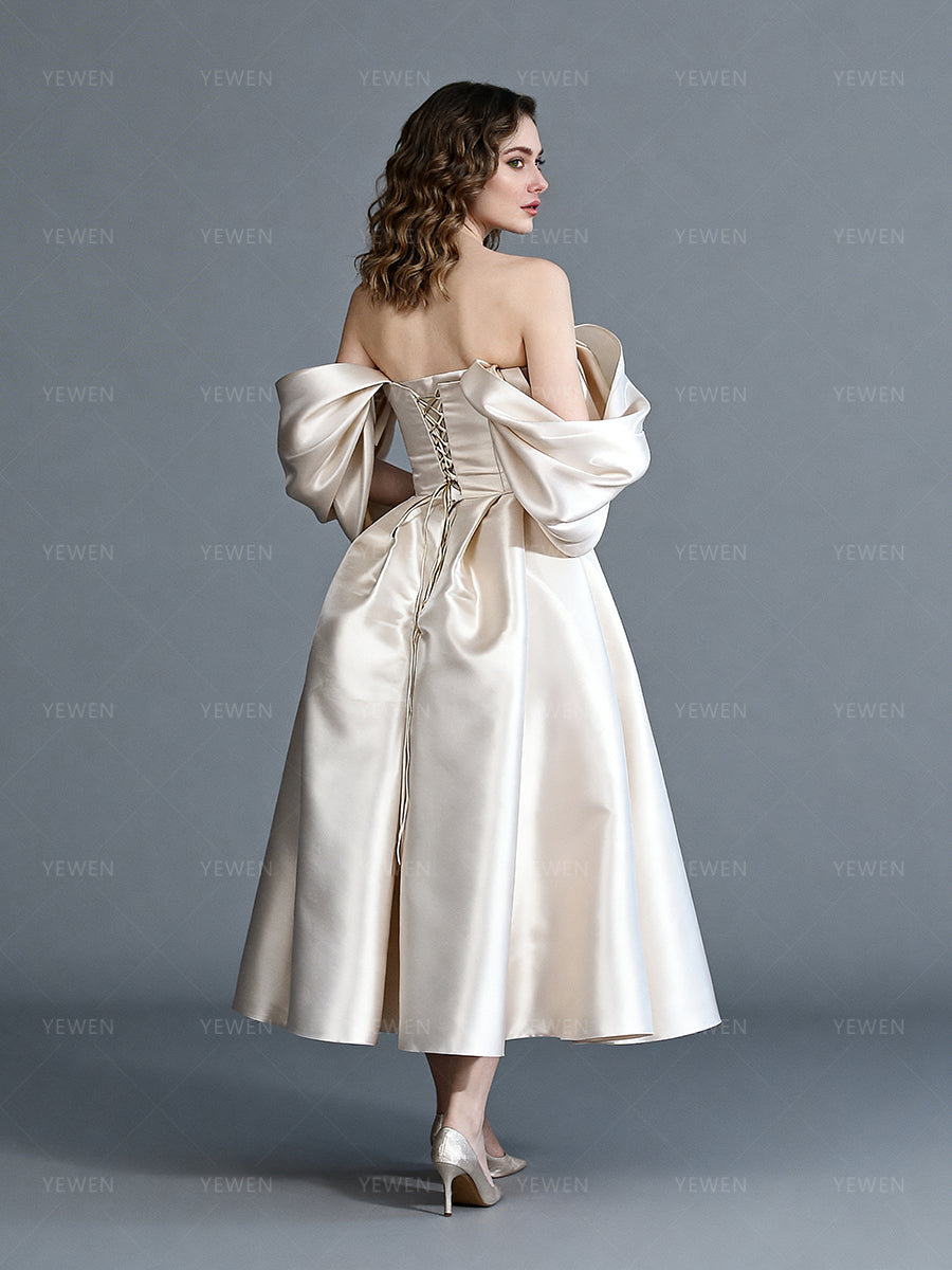Back view of a model wearing an off-shoulder satin bespoke occasion gown with a lace-up corset back and a pleated skirt.