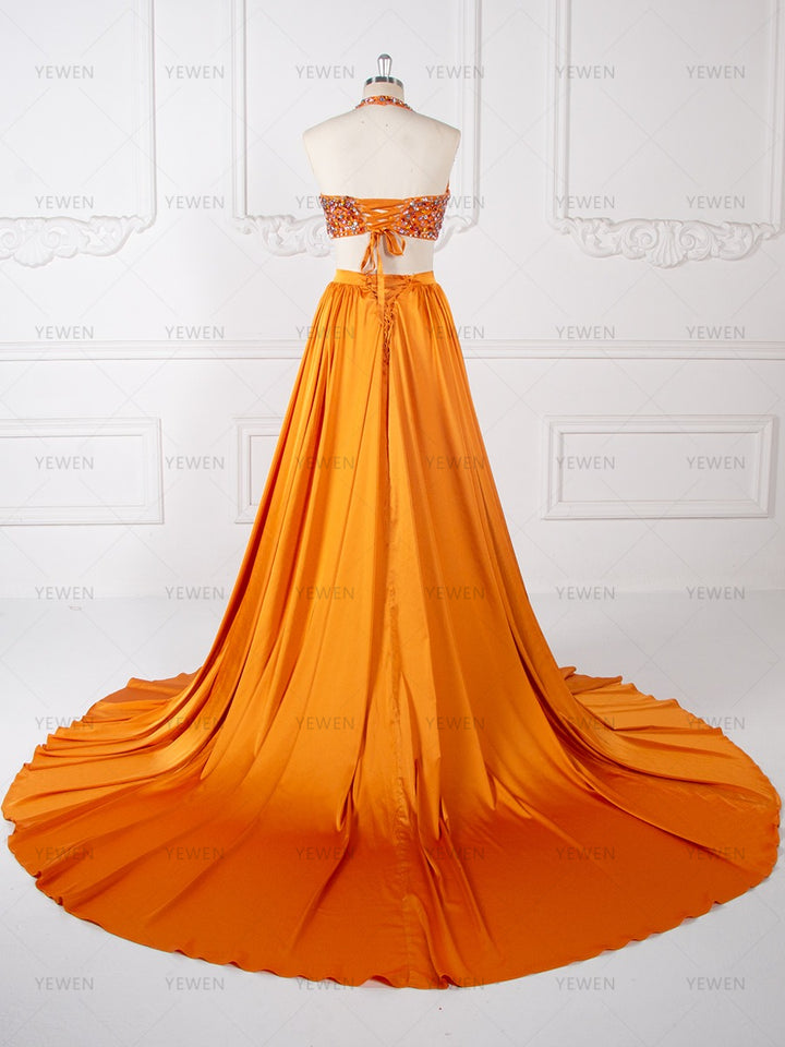 Back view of an orange beaded two-piece evening gown with a flowing pleated maxi skirt displayed on a mannequin.