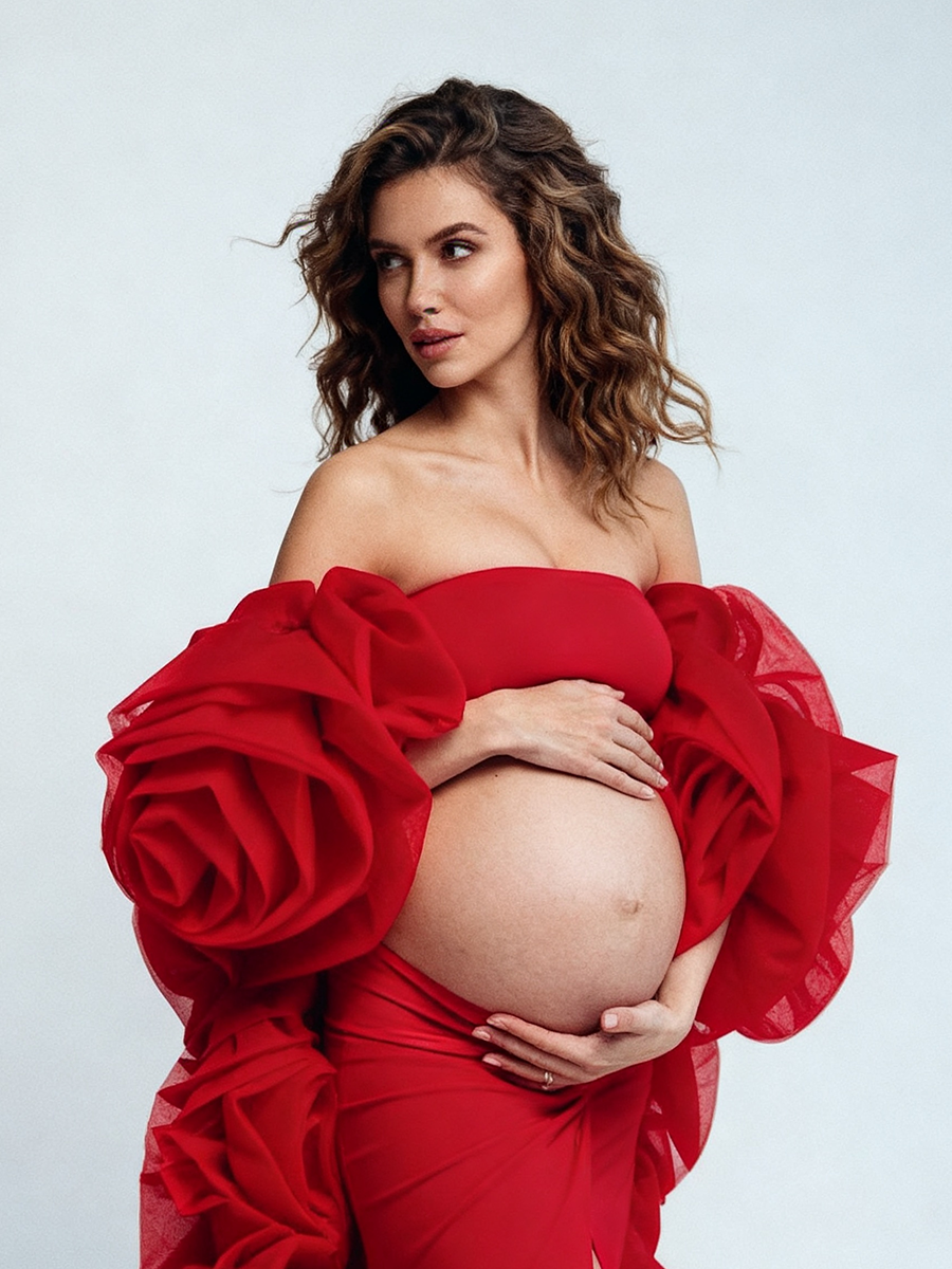 Close-up side view of a pregnant model wearing a red maternity photoshoot gown with a strapless sweetheart neckline and a dramatic oversized 3D rose flower cape.