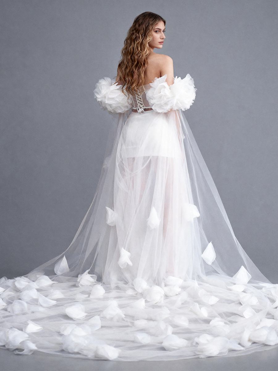 Back profile full view of a model wearing a white bridal cape with oversized 3D floral details and a sweeping sheer train.