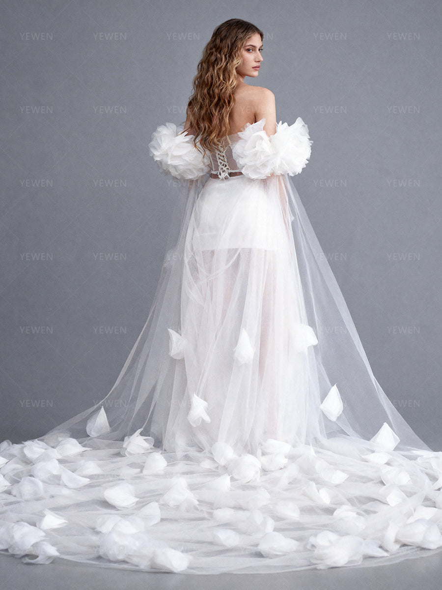 Back view of a model wearing a white bridal cape with oversized 3D floral details and a sweeping sheer train.