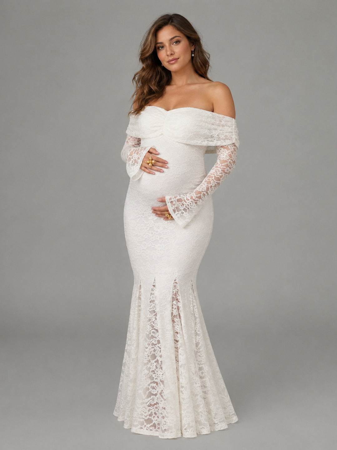 A pregnant model wearing an off-the-shoulder, long-sleeved white lace maternity gown with a fitted bodice and flared lace hem, photographed against a solid gray studio background.