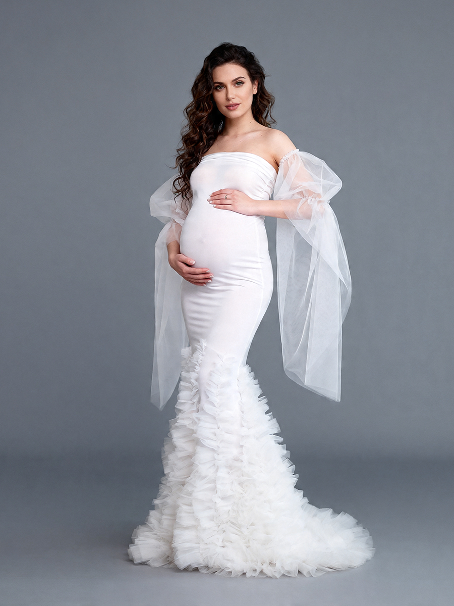 Full-body front view of a pregnant model wearing a white mermaid maternity wedding dress with a strapless sweetheart neckline sheer arm drapes and a layered ruffle train.