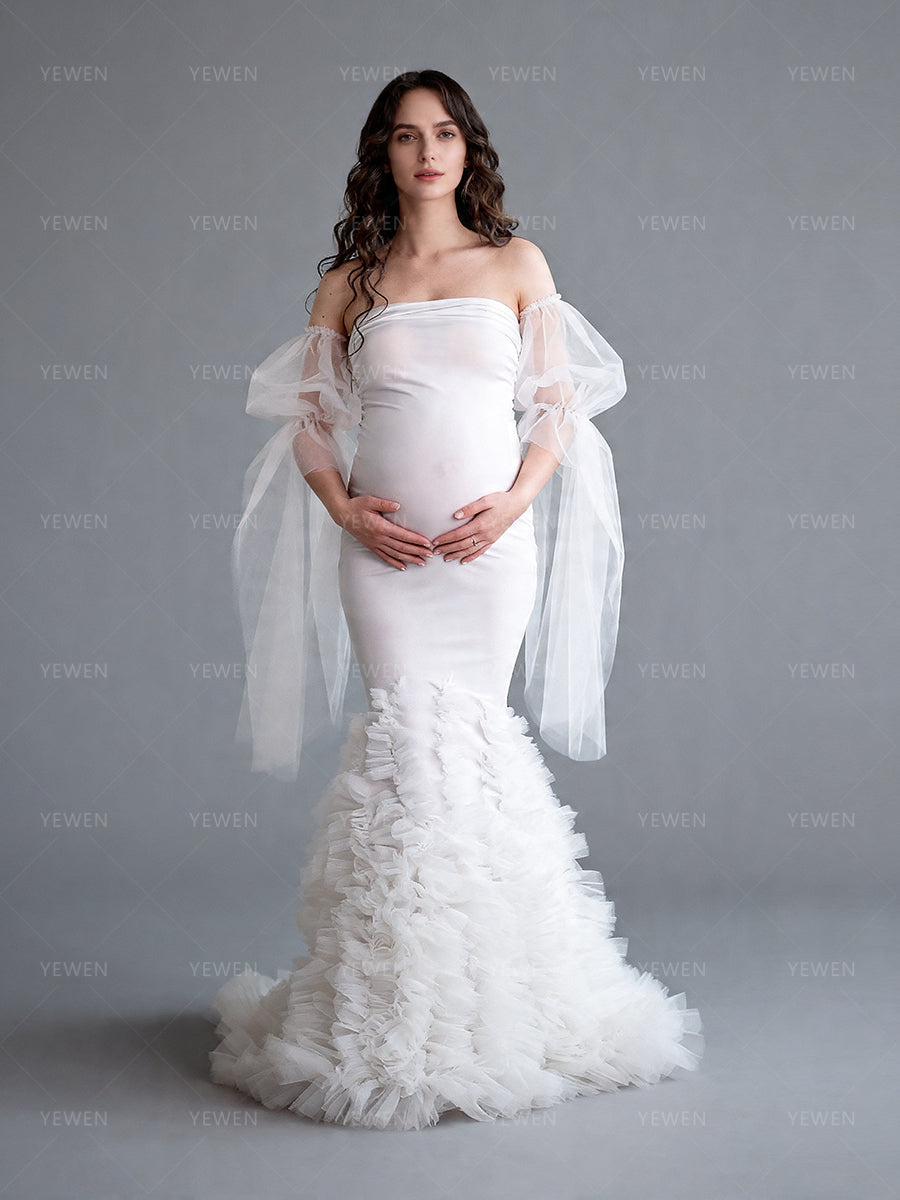 Side view of a pregnant model wearing a white mermaid maternity wedding dress with a strapless sweetheart neckline sheer arm drapes and a layered ruffle train.