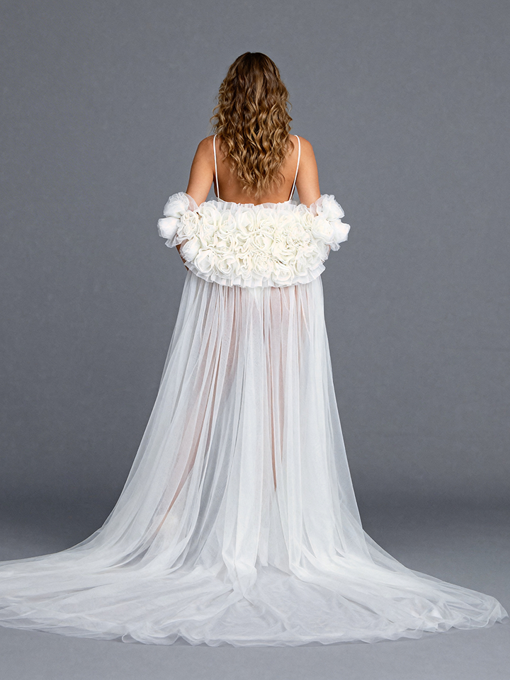 Back view of a model wearing a white tulle flower cape accessory with handmade floral details and a flowing sheer train.
